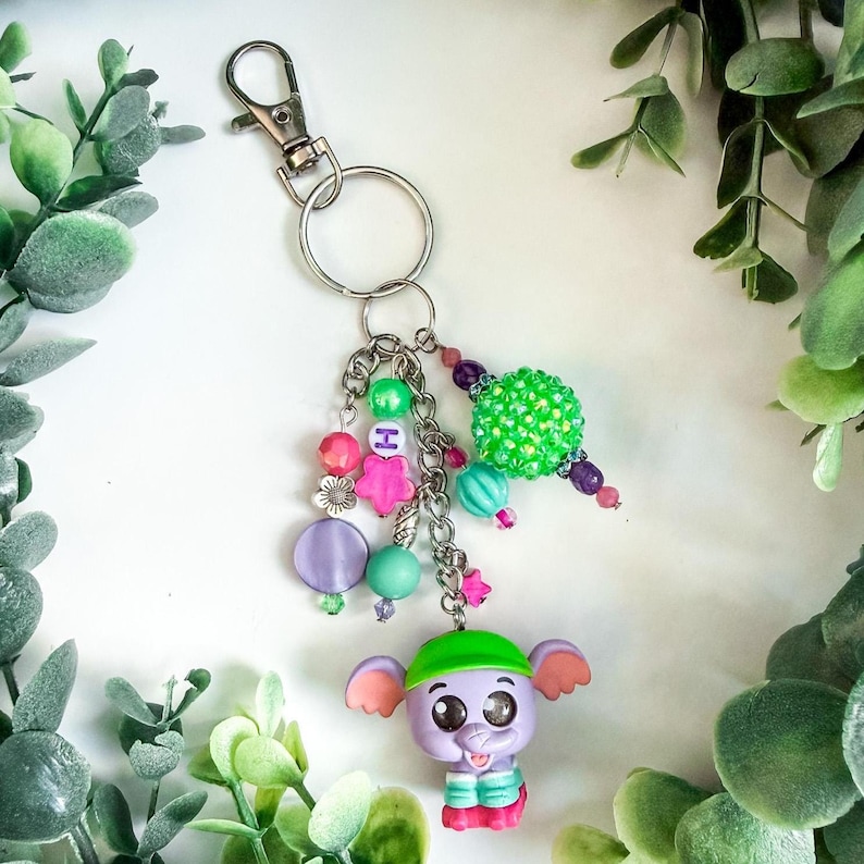 K&ouml;nnte beinhalten: Ein skurriles Schl&uuml;sselanh&auml;nger mit einer lila Elefantenfigur mit gr&uuml;nem Hut und bunten Perlenakzenten. Der Schl&uuml;sselanh&auml;nger enth&auml;lt einen silbernen Verschluss, eine Kette und verschiedene Perlen in Rosa, Gr&uuml;n und Lila.