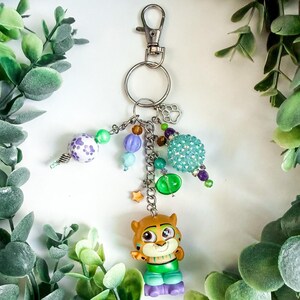 K&ouml;nnte beinhalten: Ein dekorativer Schl&uuml;sselanh&auml;nger mit einer Cartoon-Tigerfigur in gr&uuml;ner und lila Kleidung. Der Schl&uuml;sselanh&auml;nger enth&auml;lt einen silbernen Verschluss, einen Pfotenabdruck-Anh&auml;nger und verschiedene bunte Perlen in Lila-, Gr&uuml;n- und Blaut&ouml;nen.