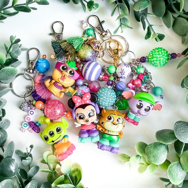 K&ouml;nnte beinhalten: Eine Sammlung bunter Schl&uuml;sselanh&auml;nger mit Cartoon-Charakteren und dekorativen Perlen. Die Schl&uuml;sselanh&auml;nger umfassen verschiedene Designs, wie einen gr&uuml;nen Alien, Minnie Mouse und einen Tiger. Jeder Schl&uuml;sselanh&auml;nger hat einen Metallverschluss.