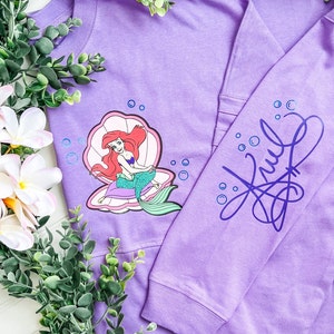 May include: Purple long-sleeved shirt with a graphic of Ariel the Little Mermaid sitting in a seashell. The shirt has a blue and purple design on the sleeve that says "Ariel".