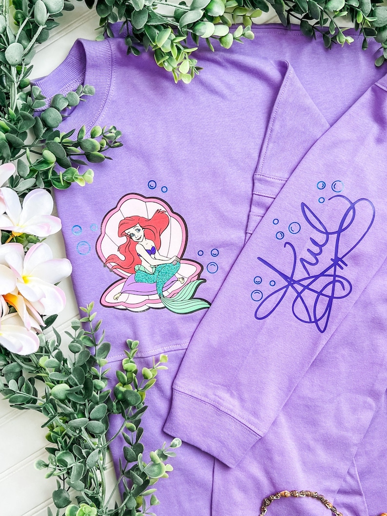 May include: Purple long-sleeved shirt with a graphic of Ariel the Little Mermaid sitting in a seashell. The shirt has a blue and purple design on the sleeve that says "Ariel".