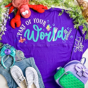 May include: Purple long-sleeved shirt with the text "Part of Your World" in teal glitter. The shirt features a small image of Ariel and Sebastian. The shirt is laid out on a white surface with a pair of blue jeans, a pair of white shoes, a pair of mermaid ears, and a purple and green backpack.