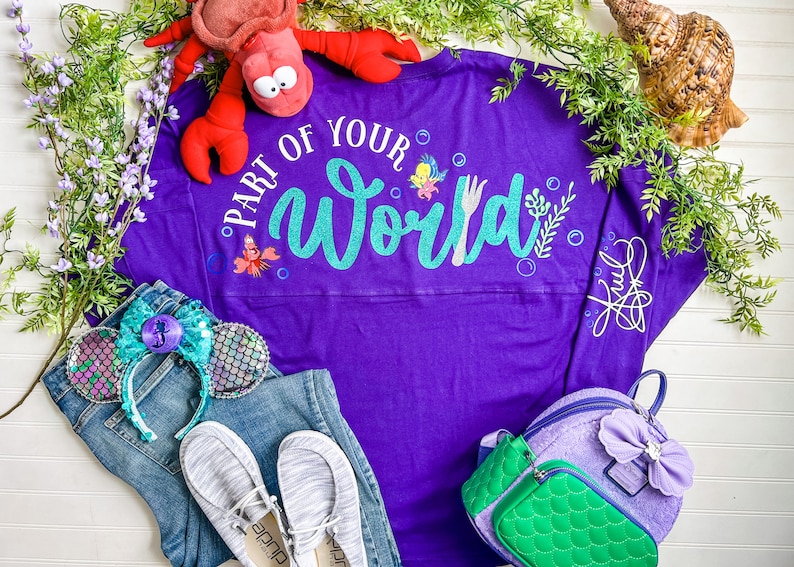 May include: Purple long-sleeved shirt with the text "Part of Your World" in teal glitter. The shirt features a small image of Ariel and Sebastian. The shirt is laid out on a white surface with a pair of blue jeans, a pair of white shoes, a pair of mermaid ears, and a purple and green backpack.