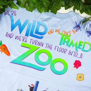 Puede incluir: Camiseta azul claro con las palabras "WE'RE WILD AND WE CAN'T BE TAMED" en un dise&ntilde;o degradado colorido. La camiseta tambi&eacute;n presenta la palabra "ZOO" y gr&aacute;ficos de dibujos animados de una zanahoria, un helado y una placa de polic&iacute;a.