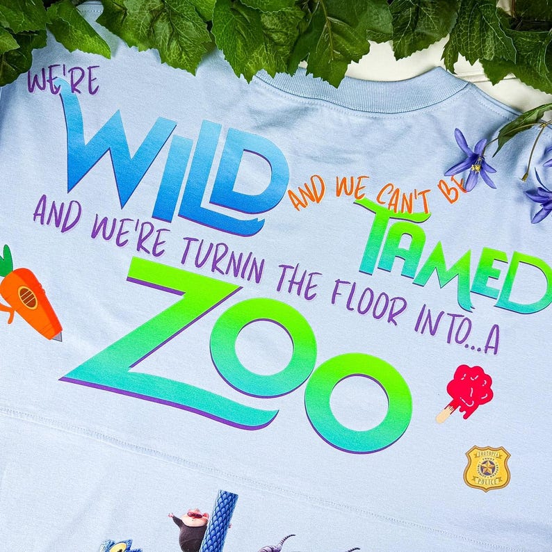 Puede incluir: Camiseta azul claro con las palabras "WE'RE WILD AND WE CAN'T BE TAMED" en un dise&ntilde;o degradado colorido. La camiseta tambi&eacute;n presenta la palabra "ZOO" y gr&aacute;ficos de dibujos animados de una zanahoria, un helado y una placa de polic&iacute;a.