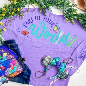 May include: Purple long-sleeved shirt with the text "Part of Your World" in teal glitter with a fork and a small cartoon crab. The shirt has a teal and silver glitter design on the sleeve.