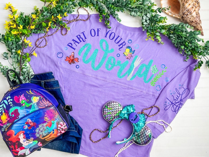 May include: Purple long-sleeved shirt with the text "Part of Your World" in teal glitter with a fork and a small cartoon crab. The shirt has a teal and silver glitter design on the sleeve.