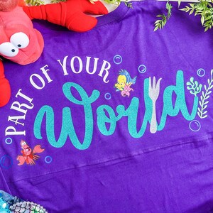 May include: Purple long-sleeved shirt with the text "Part of Your World" in white and teal glitter. The shirt features a fork, a seahorse, and a cartoon fish.