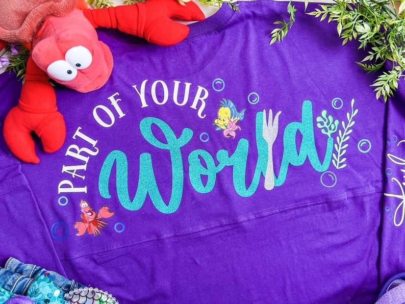 May include: Purple long-sleeved shirt with the text "Part of Your World" in white and teal glitter. The shirt features a fork, a seahorse, and a cartoon fish.