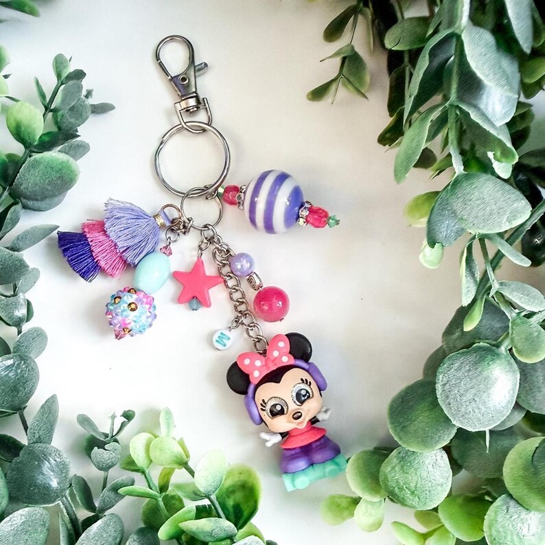 K&ouml;nnte beinhalten: Ein dekorativer Schl&uuml;sselanh&auml;nger mit einer Minnie-Mouse-Figur, einem rosa Sternanh&auml;nger und verschiedenen Perlen in Rosa, Lila und Blau. Der Schl&uuml;sselanh&auml;nger hat einen silbernen Verschluss und einen Quasten.