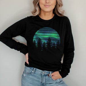 Pode incluir: Camisola preta de manga comprida com um gráfico de estilo retro de uma aurora boreal verde e azul sobre uma silhueta de uma floresta. O gráfico está em um círculo com um fundo estrelado.