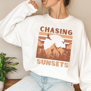 May include: White sweatshirt with a graphic design of mountains, a sun, and birds. The text "CHASING SUNSETS" is printed on the sweatshirt.