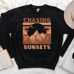 May include: Black sweatshirt with a brown graphic design featuring mountains, a sunset, and birds. The text "CHASING SUNSETS" is printed on the sweatshirt.