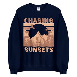 May include: Navy blue sweatshirt with a brown graphic of mountains, a sunset, and birds flying. The text "CHASING SUNSETS" is printed in brown on the sweatshirt.