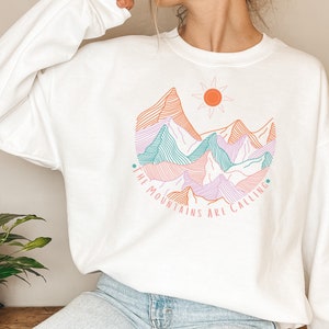 Könnte beinhalten: Weißer Pullover mit einem Motiv von Bergen, einer Sonne und dem Text "The Mountains Are Calling".