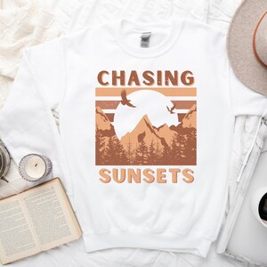 May include: White sweatshirt with a brown graphic design that says "CHASING SUNSETS" with a mountain range and two birds flying in the sky.