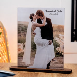 May include: A wedding photo of a couple kissing in a field with tall grass. The photo is in a clear acrylic frame with a wooden base. The frame has the text "Samanta & John 2022 11 11" printed on it.