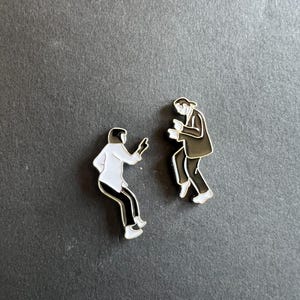 Könnte beinhalten: Zwei Emaille-Pins, die eine schwarz-weiße Silhouette von zwei tanzenden Personen zeigen. Die Person links trägt ein weißes Hemd und eine schwarze Hose, während die Person rechts einen schwarzen Anzug trägt.