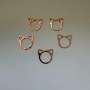 Könnte beinhalten: Fünf kupferfarbene Metallringe, die jeweils oben eine ausgeprägte Katzenohr-Silhouette aufweisen. Die Ringe haben eine glatte, metallische Oberfläche und ein kreisförmiges Banddesign und sind in einer Fünfeckform auf neutralem Grund angeordnet.