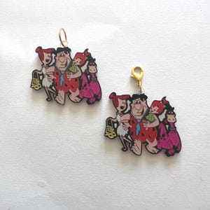 Könnte beinhalten: Zwei Cartoon-Charakter-Anhänger mit der Familie Feuerstein. Jeder Anhänger zeigt Fred, Wilma, Pebbles und Betty. Die Anhänger sind farbenfroh mit goldfarbenen Sprungringen zum Aufhängen. Der Hintergrund ist weiß.