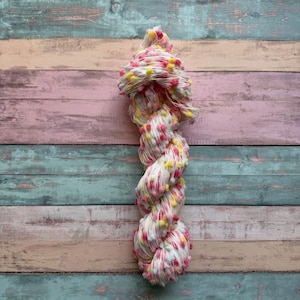 Könnte beinhalten: Ein Knäuel weißer Wolle mit rosa und gelben Pompons. Die Wolle ist verdreht und geknotet und vor einem Hintergrund aus pastellfarbenen Holzbrettern platziert. Dieses Garn eignet sich für Strick- und Häkelprojekte.
