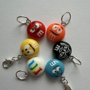 Könnte beinhalten: Sechs bunte M&M's-Anhänger mit silberfarbenen Karabinerverschlüssen und Schlüsselringen. Die Anhänger sind rot, orange, gelb, schwarz, weiß und blau, jeder mit einem anderen Gesichtsausdruck und dem Buchstaben "m".
