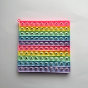Könnte beinhalten: Ein farbenfrohes quadratisches Zappelspielzeug mit einem Regenbogenverlauf und nummerierten Blasen von 1 bis 100. Das Spielzeug hat einen rosa Rand und eine kleine Schlaufe oben. Die Blasen sind in einem Raster angeordnet.
