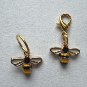 Könnte beinhalten: Zwei goldfarbene Bienen-Anhänger. Jeder Anhänger hat einen gelb-schwarzen Körper mit weißen Flügeln. Einer hat einen kleinen Ring, der andere einen Verschluss. Geeignet für Schmuckherstellung oder Bastelprojekte.