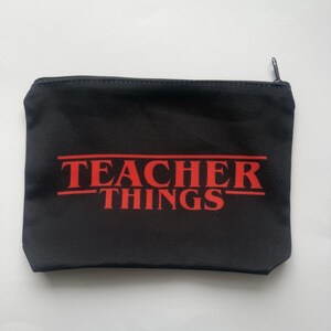 Könnte beinhalten: Schwarze, rechteckige Reißverschlusstasche mit rotem Aufdruck "TEACHER THINGS". Die Tasche besteht aus einem dunklen Stoff und ist für kleine Gegenstände konzipiert. Der Reißverschluss ist schwarz.