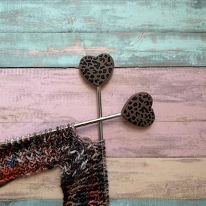 Könnte beinhalten: Zwei Stricknadelstopper mit herzförmigen Oberteilen mit Leopardenmuster liegen auf einer farbenfrohen Holzoberfläche. Die Nadeln befinden sich in der Nähe eines teilweise gestrickten Garnstücks in Rot-, Braun- und Weißtönen.
