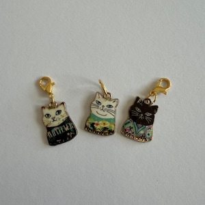 Könnte beinhalten: Drei katzenförmige Anhänger mit goldfarbenen Verschlüssen. Jeder Anhänger zeigt ein anderes Katzendesign mit bunten Mustern und Details. Die Katzen haben verschiedene Ausdrücke und tragen dekorative Outfits. Die Anhänger sind wahrscheinlich für Schmuck oder Bastelarbeiten.