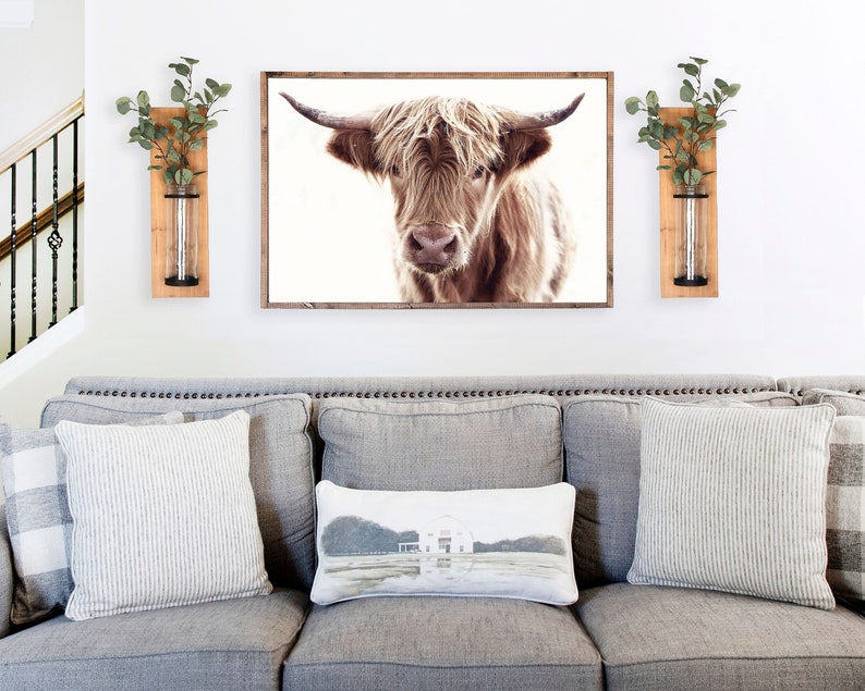 May include: A gray couch with three pillows, one with a farmhouse scene, in front of a white wall with a framed print of a brown Highland cow and two wooden wall vases with greenery.