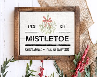 May include: A rustic wooden sign with a white background and black lettering. The sign reads "Fresh Cut Mistletoe 25 Cents a Bundle Kisses are Always Free". A green and red mistletoe sprig with a red bow is pictured above the text.