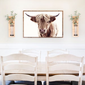 May include: A white dining room with a wooden table and white chairs. A framed print of a brown Highland cow hangs on the wall above the table. Two wooden wall vases with greenery are on either side of the print.