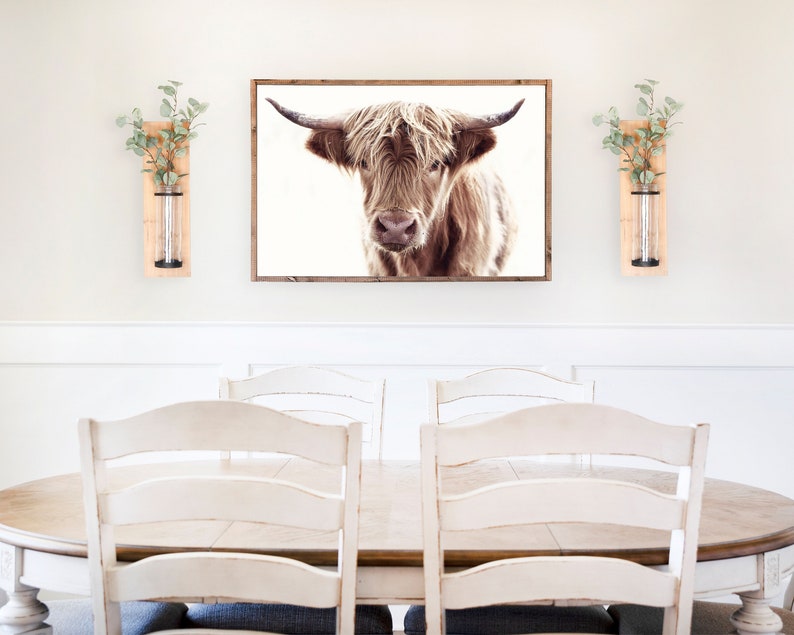 May include: A white dining room with a wooden table and white chairs. A framed print of a brown Highland cow hangs on the wall above the table. Two wooden wall vases with greenery are on either side of the print.