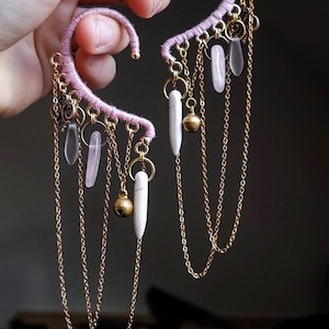 Może przedstawiać: Para nausznic z różową, owiniętą ramą, ozdobionych złotymi łańcuszkami, małymi złotymi koralikami i zwisającymi białymi i przezroczystymi akcentami przypominającymi kryształ. Ciemne tło.