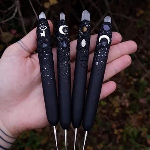 Peut inclure: Quatre stylos noirs avec des dessus décoratifs ornés de cristaux et de symboles célestes. Chaque stylo a une pointe argentée et est tenu dans une main sur un fond naturel flou. Les stylos ont des motifs uniques, dont une clé, un croissant de lune et des étoiles.