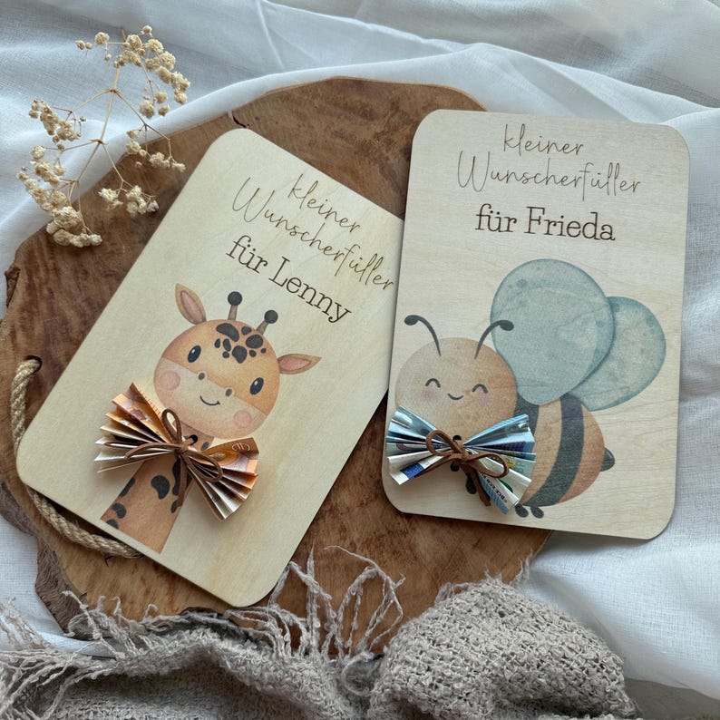 May include: Two wooden gift cards with illustrations and text. One card features a giraffe with a paper bow, the other a bee with a paper bow. The text on the cards reads "Kleiner Wunscherf&uuml;ller f&uuml;r Lenny" and "Kleiner Wunscherf&uuml;ller f&uuml;r Frieda".