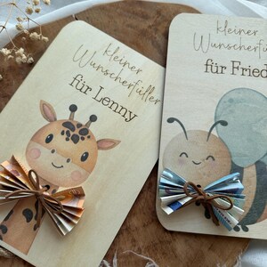 May include: Two wooden cards with illustrations and text. One card features a giraffe with a bow made of folded paper money. The other card has a bee with a similar bow. The text on the cards is in German.
