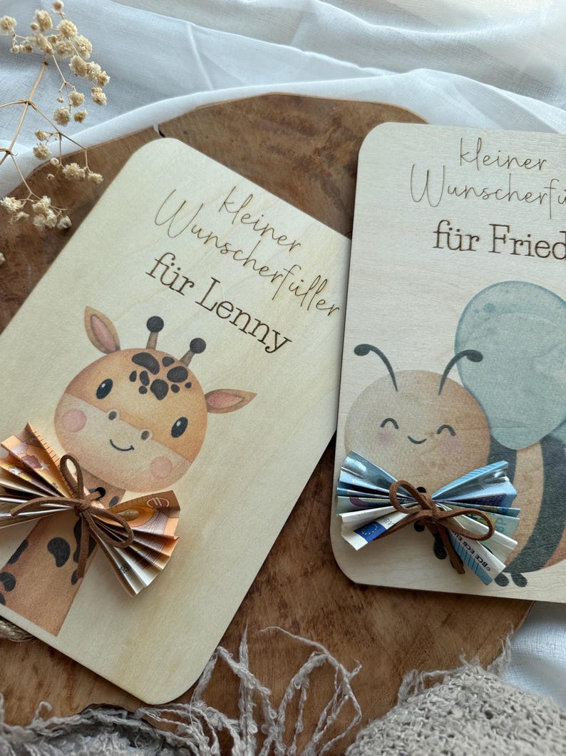 May include: Two wooden cards with illustrations and text. One card features a giraffe with a bow made of folded paper money. The other card has a bee with a similar bow. The text on the cards is in German.