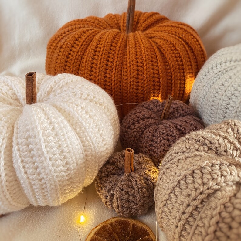 May include: A collection of crocheted pumpkins in various colors and sizes, including orange, white, brown, and beige. The pumpkins are arranged on a white surface with string lights.