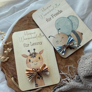 May include: Two wooden cards with illustrations and text. One card features a giraffe, the other a bee with blue wings. Both cards have folded paper bows and the text "kleiner Wunscherf&uuml;ller f&uuml;r Lenny" and "kleiner Wunscherf&uuml;ller f&uuml;r Frieda".