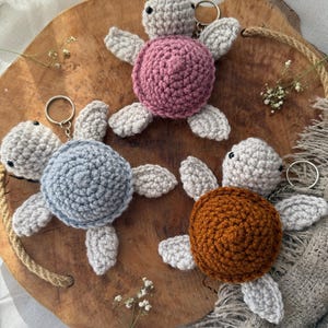 Könnte beinhalten: Drei gehäkelte Schildkröten-Schlüsselanhänger in Rosa, Grau und Braun. Die Schlüsselanhänger sind aus weichem Garn gefertigt und haben ein süßes, verspieltes Design.