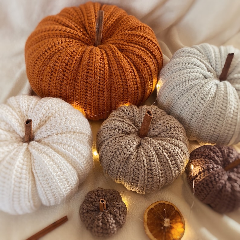 May include: Five crocheted pumpkins in various sizes and colors, including orange, white, beige, and brown. The pumpkins are arranged on a white surface with string lights and cinnamon sticks.