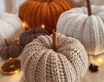 May include: A collection of crocheted pumpkins in various sizes and colors, including orange, white, brown, and beige. The pumpkins are arranged on a white surface with string lights and decorative elements.
