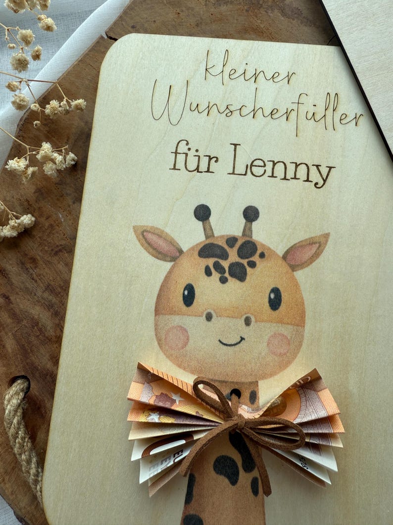 May include: A wooden plaque with a giraffe illustration and the text "kleiner Wunscherf&uuml;ller f&uuml;r Lenny". The giraffe has brown spots and a bow made of folded banknotes.