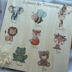 May include: Wooden board featuring watercolor illustrations of various animals, including an elephant, bee, lion, fox, frog, bear, giraffe, and deer. The text "Auswahl der Tiermotive" is printed at the top.