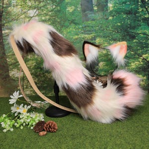 Puede incluir: Un conjunto de cola y orejas de gato de piel sintética. La cola es marrón, blanca y rosa, con un cinturón beige. Las orejas son negras, blancas y rosas. Para cosplay o disfraces.