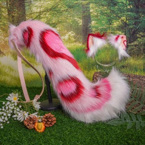 May include: Pink and red fluffy tail with a white tip, attached to a brown leather strap. The tail is a costume accessory for a cat or other animal.