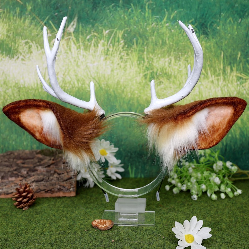 May include: A pair of deer antlers and ears headband. The antlers are white, and the ears are brown with white and tan accents. The headband is clear plastic. The background is a blurred green with flowers.